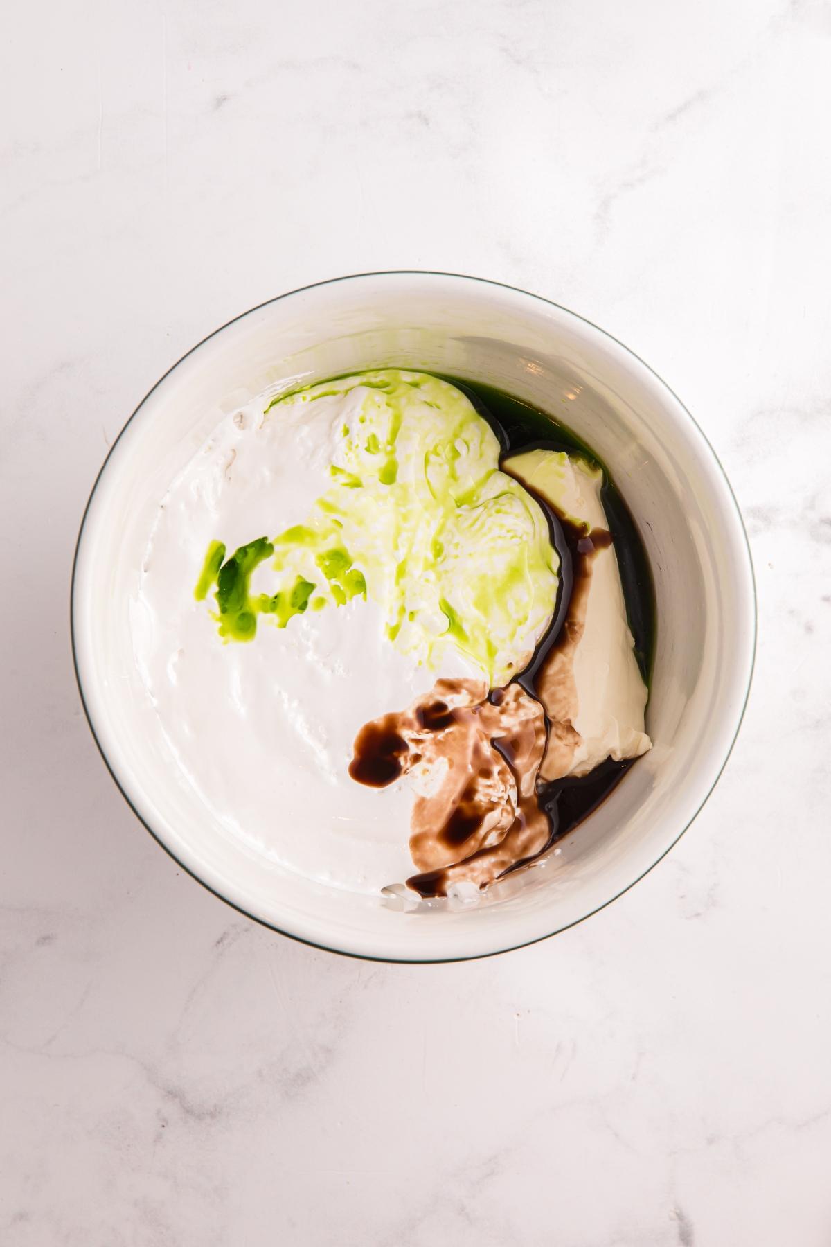 A mixing bowl with cream, green food coloring, vanilla, and cream cheese on a white marble surface.