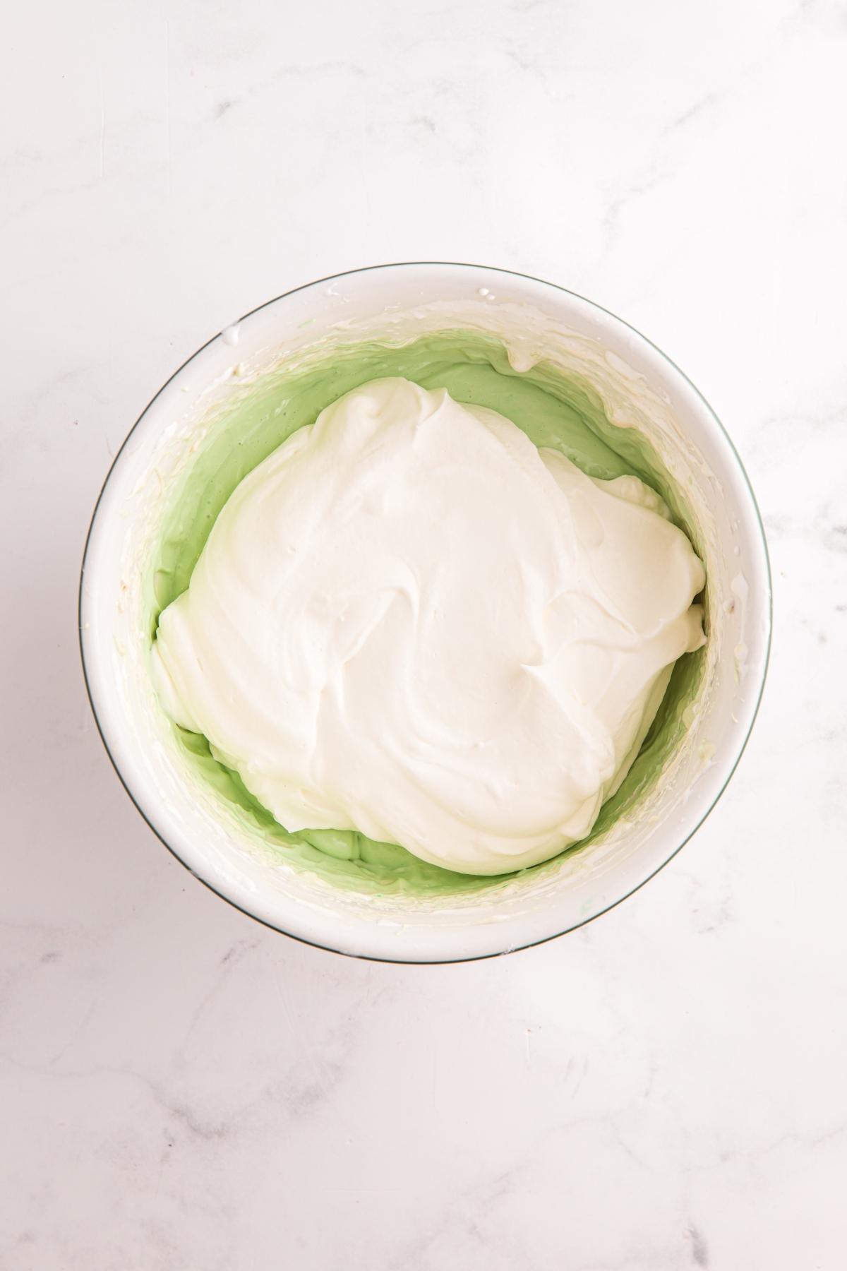 A bowl containing green pudding mixture topped with a layer of fluffy white whipped cream.