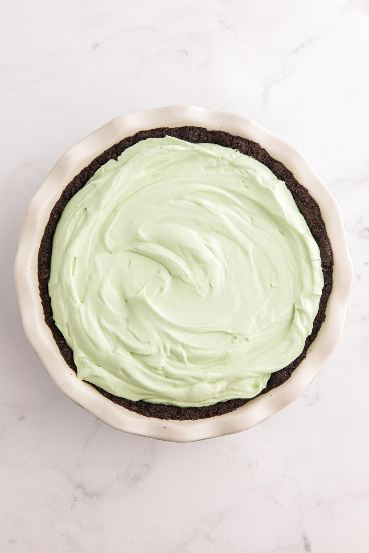 A chocolate pie crust filled with light green mint cream, in a white pie dish on a marble surface.