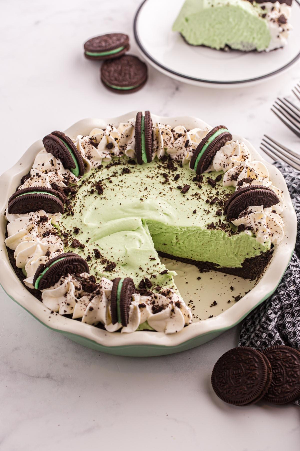 A grasshoppe pie with whipped cream, Oreo cookies, and cookie crumbs, with one slice served on a plate.