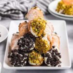 A plate of homemade cannoli with chocolate chips and chopped pistachios, surrounded by bowls of toppings.
