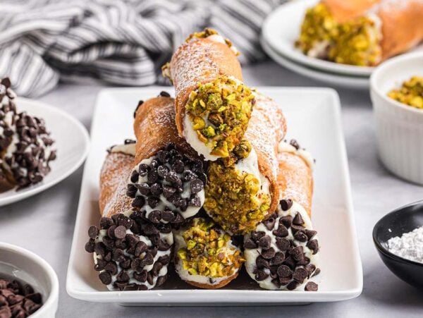 A plate of homemade cannoli with chocolate chips and chopped pistachios, surrounded by bowls of toppings.