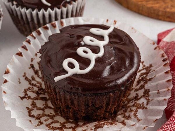 Chocolate cupcake with glossy frosting and a white icing swirl, sitting in an open paper liner next to a red napkin—just like classic Homemade Hostess Cupcakes.