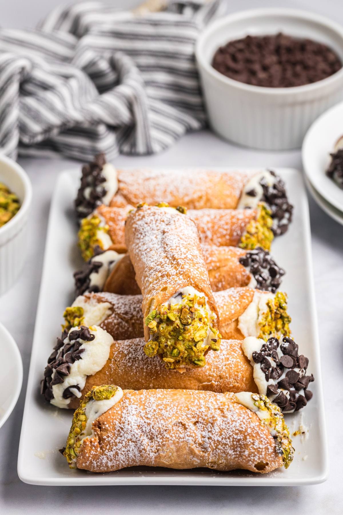A plate of cannoli filled with cream, dipped in chocolate chips and pistachios, dusted with powdered sugar.