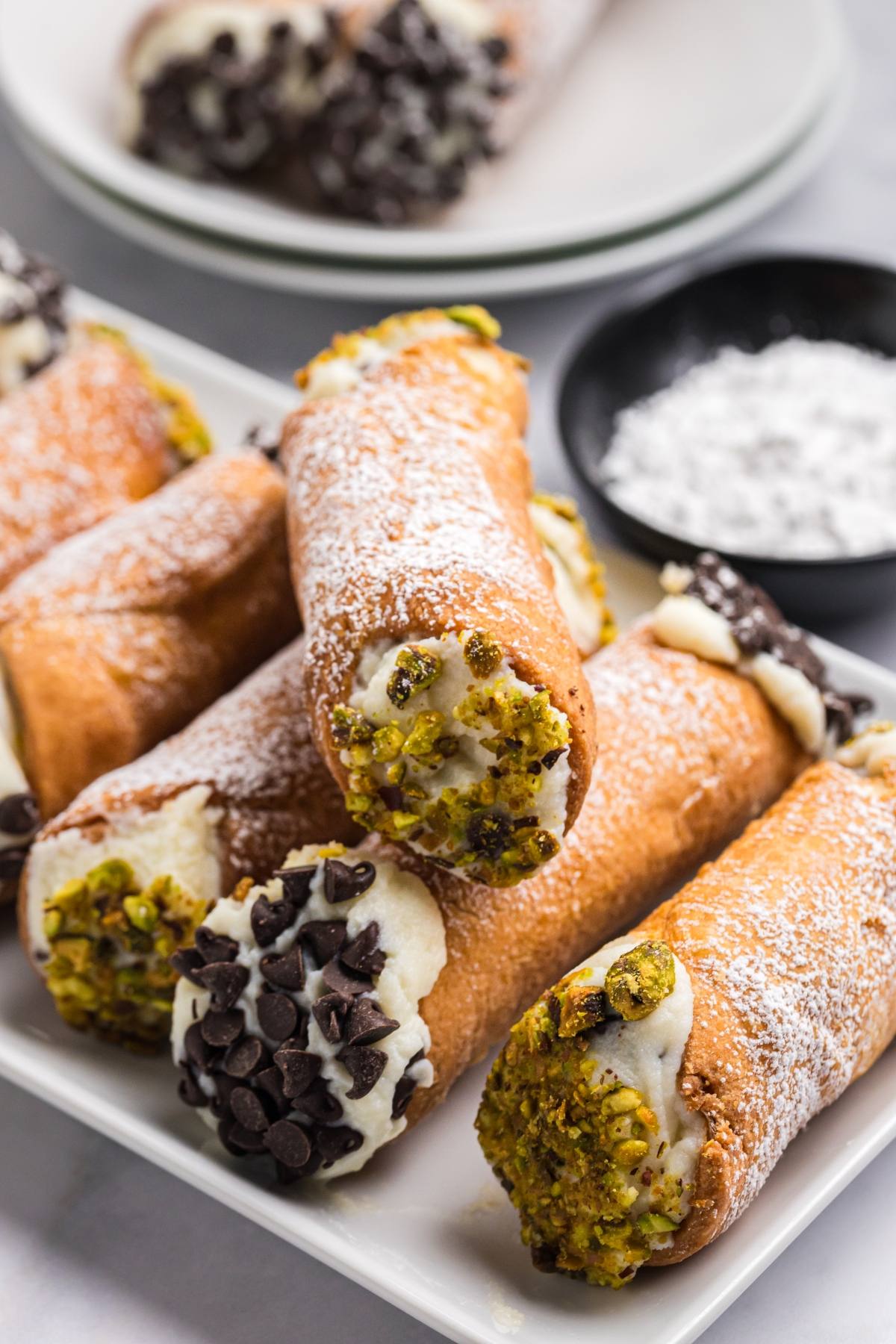 A plate of cannoli filled with cream, ends dipped in chocolate chips and crushed pistachios, dusted with powdered sugar.