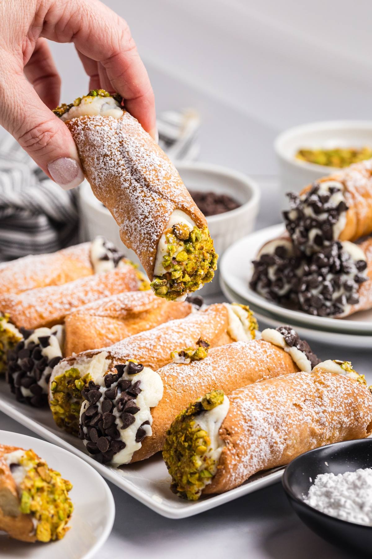 A hand holds a cannoli topped with pistachios above a plate of assorted cannoli with powdered sugar.
