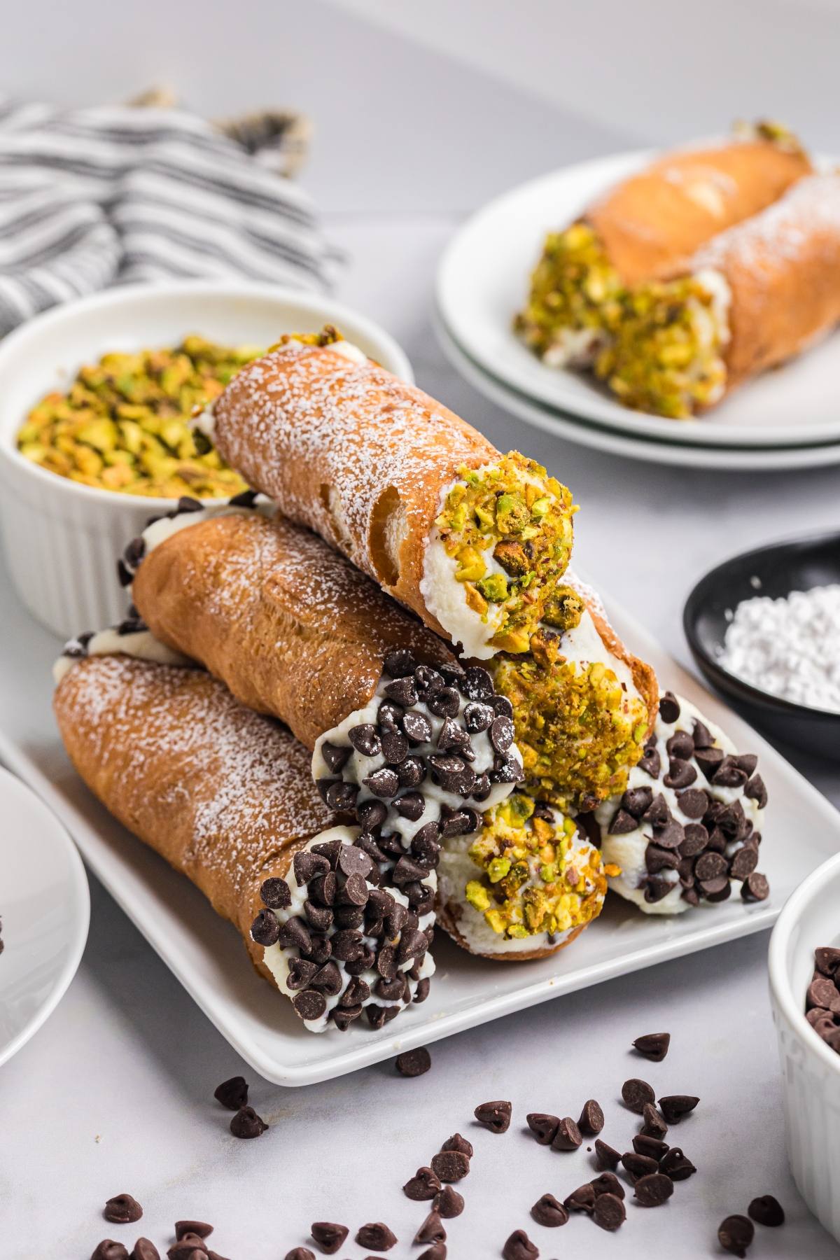 A plate of cannoli filled with cream and topped with chocolate chips or crushed pistachios, dusted with powdered sugar.