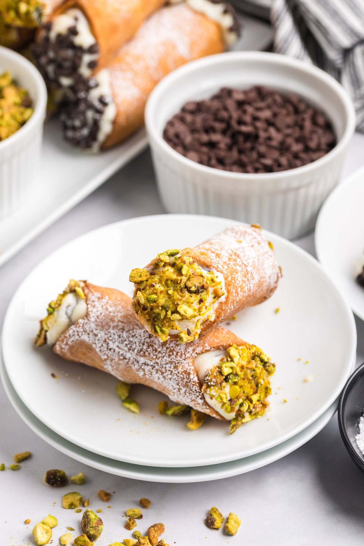 Two cannoli with pistachio pieces on a white plate, surrounded by bowls of chocolate chips and more cannoli.