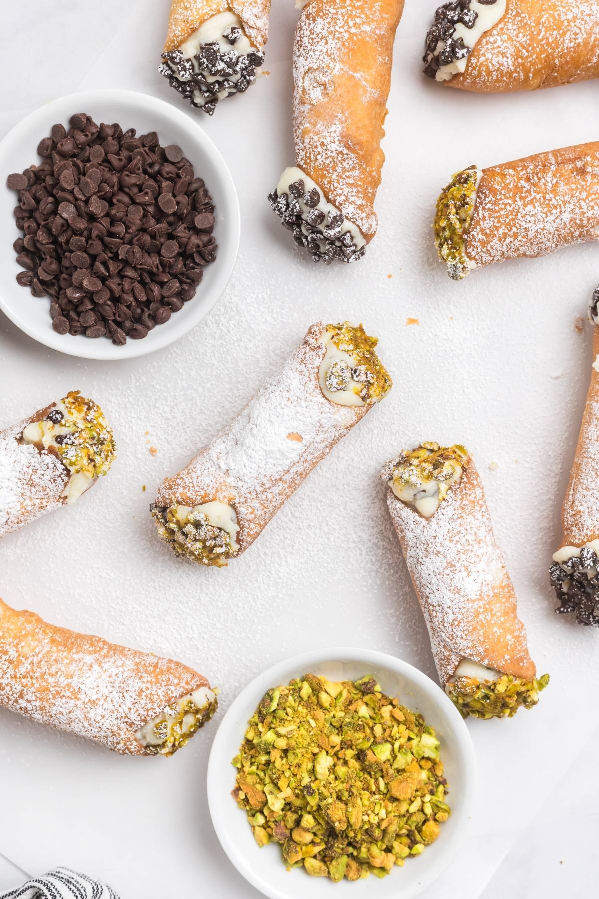 Cannoli with powdered sugar, chocolate chips, and pistachios on a white surface, with bowls of chips and nuts.