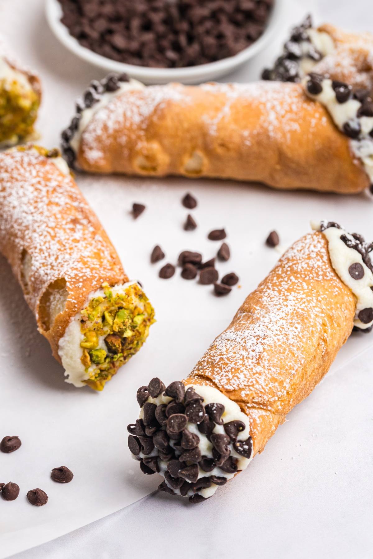 Three cannoli with ricotta filling, topped with chocolate chips and pistachios, dusted with powdered sugar.
