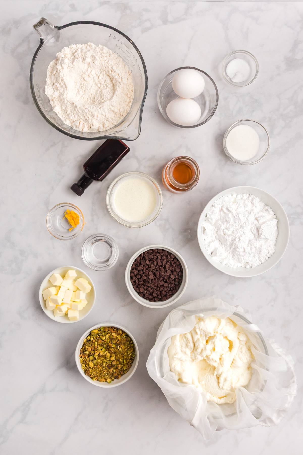 Assorted baking ingredients in bowls on a marble surface, including eggs, flour, nuts, chocolate chips, and mascarpone.