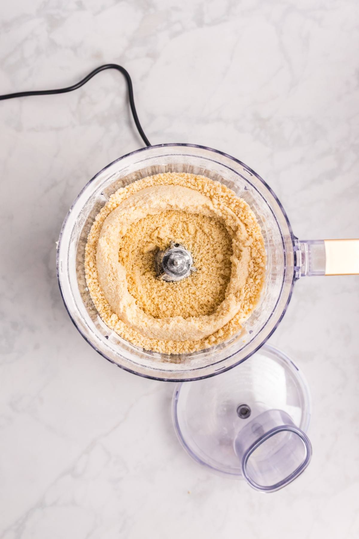 Top view of a food processor with ground almond flour, lid off, on a light marble countertop.
