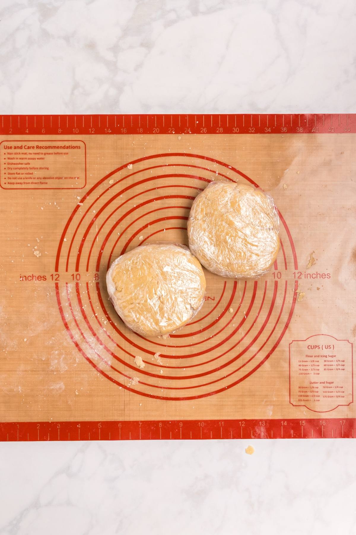 Two discs of dough wrapped in plastic wrap on a red silicone baking mat with measurement guides.