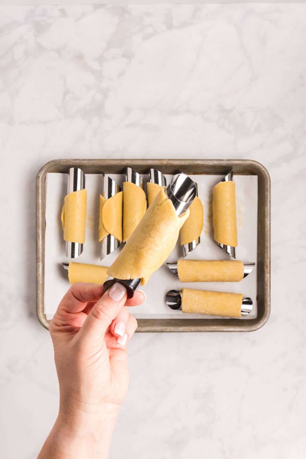 A hand holds an unbaked pastry wrapped around a metal tube, with more on a tray in the background.