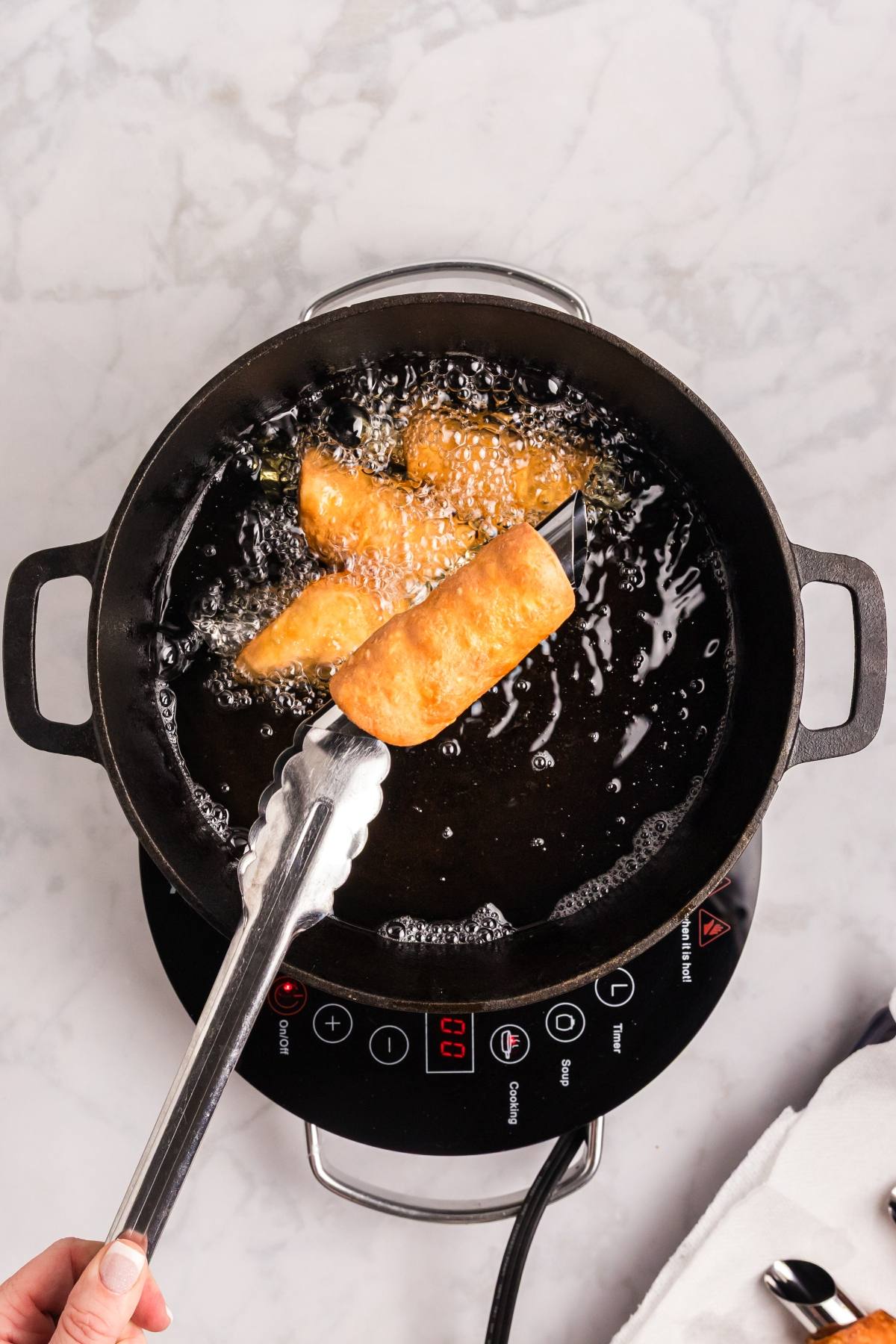 A hand uses tongs to fry golden pastries in a pot of hot oil on an electric stove.