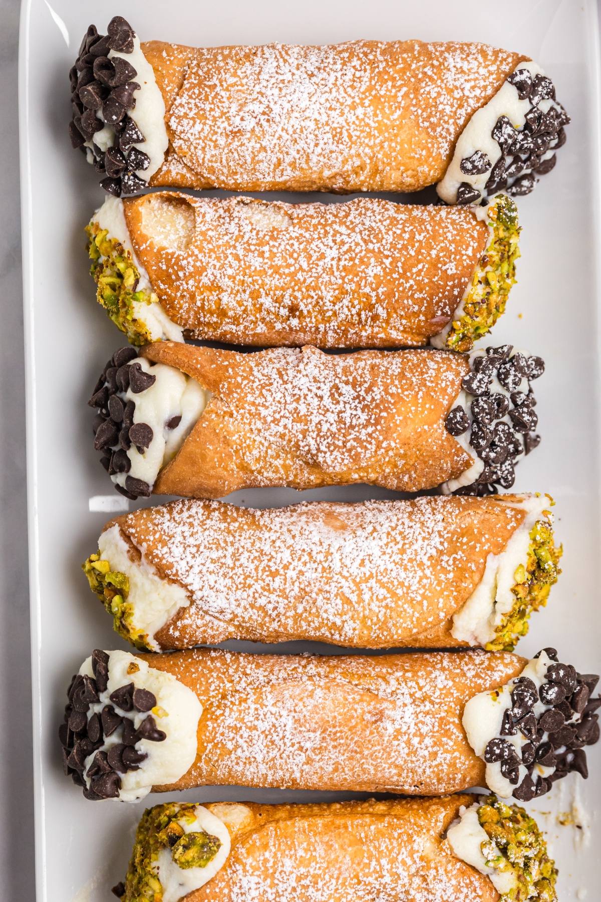 Six cannoli with ricotta filling, dusted with powdered sugar, topped with chocolate chips and chopped pistachios.