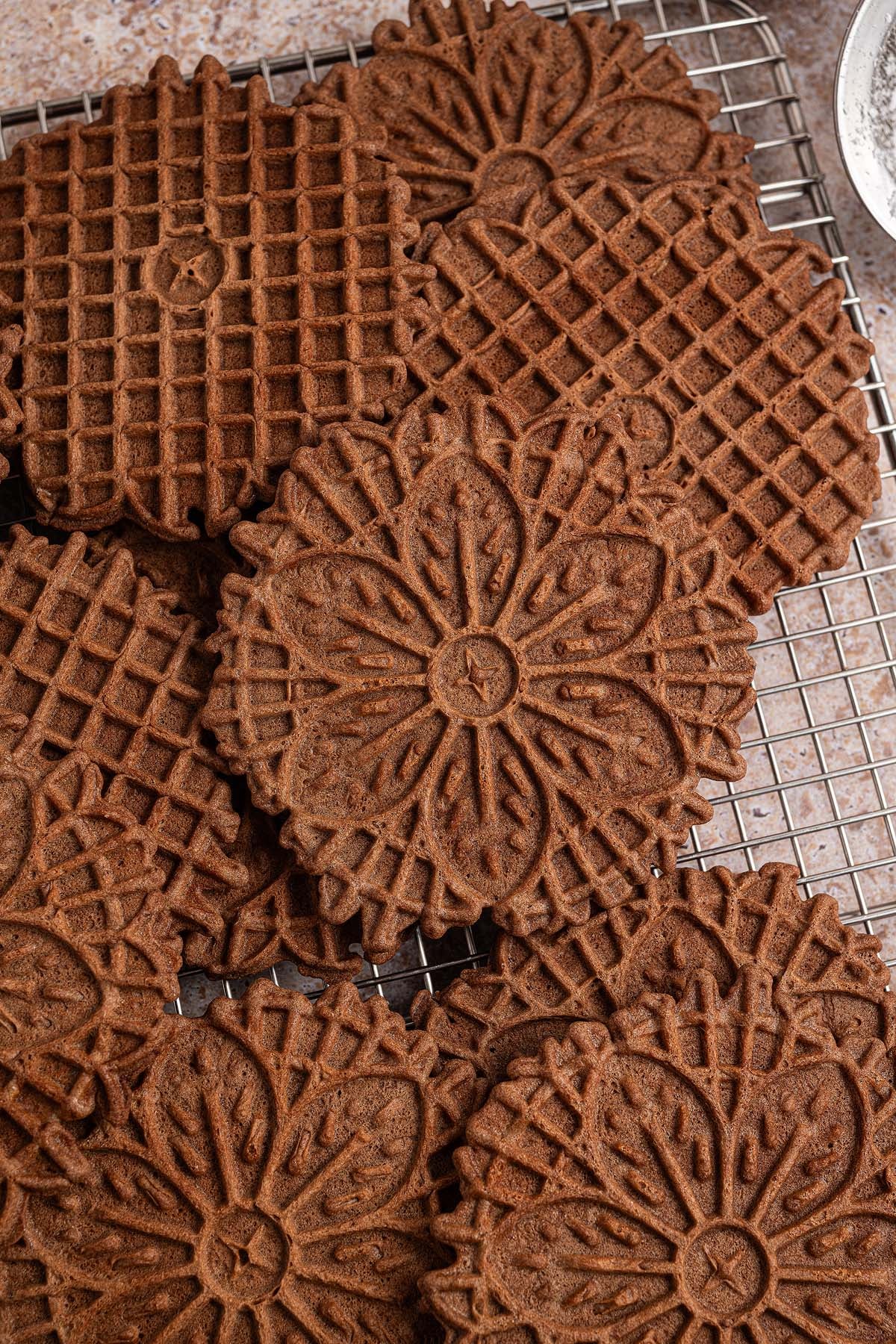 Chocolate pizzelle cookies with intricate patterns are stacked on a cooling rack, showcasing the delicate beauty of classic chocolate pizzelle.