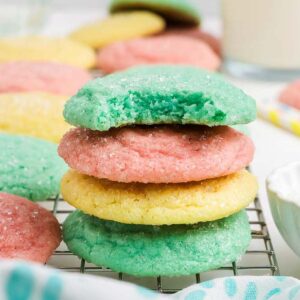 A stack of colorful Kool-Aid Cookies, with a green cookie on top showing a bite taken out, rests on a cooling rack.