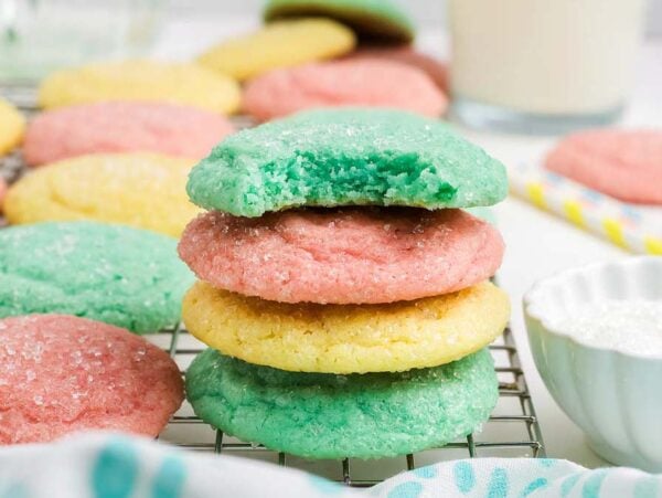 A stack of colorful Kool-Aid Cookies, with a green cookie on top showing a bite taken out, rests on a cooling rack.