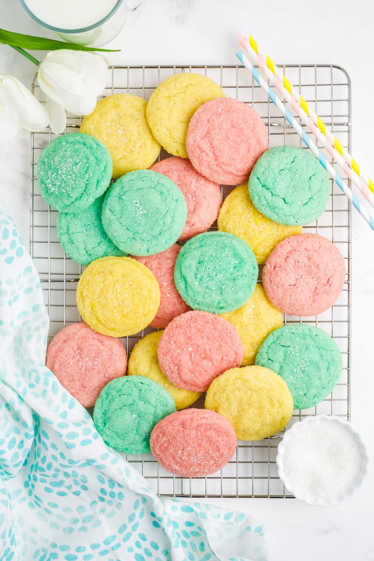 Colorful sugar cookies in pink, yellow, and green on a cooling rack with a glass of milk and pastel straws.