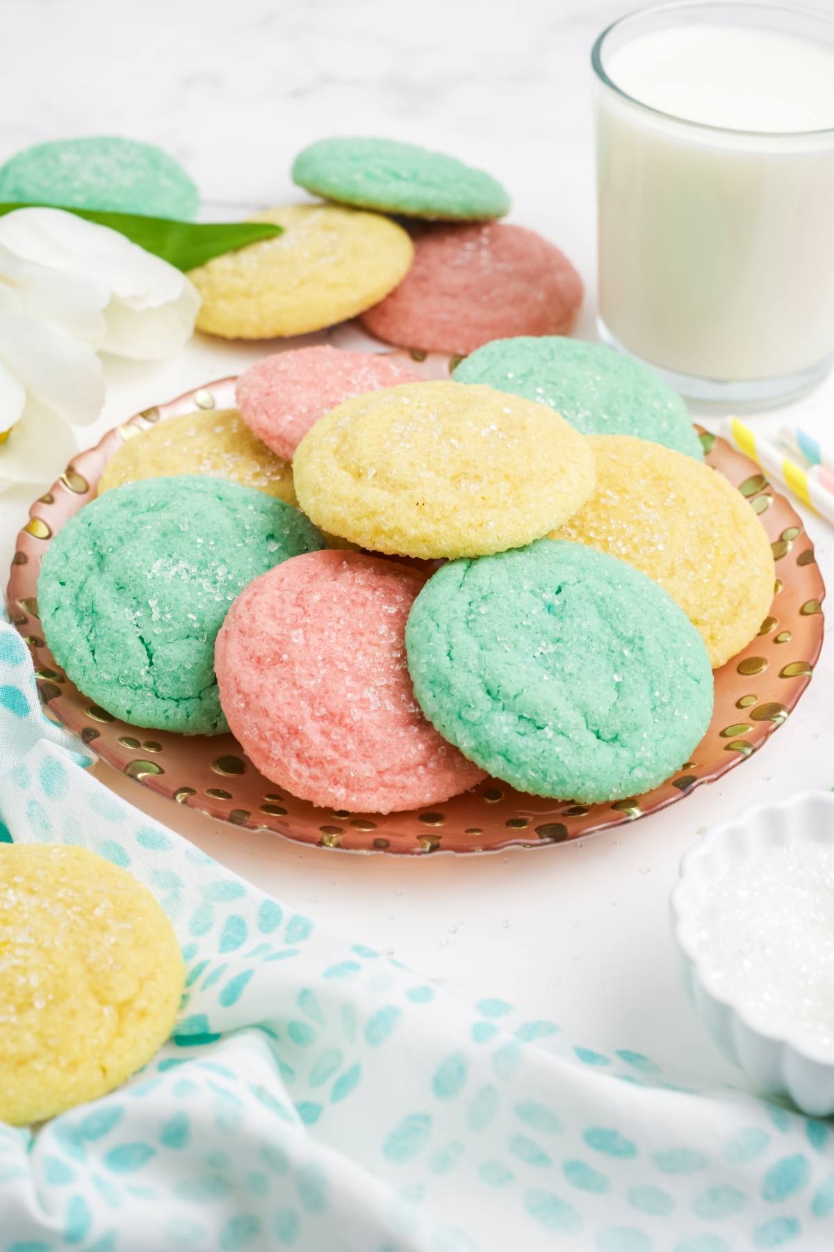 A plate of pastel-colored sugar Kool-Aid cookies with a glass of milk and a white flower nearby.