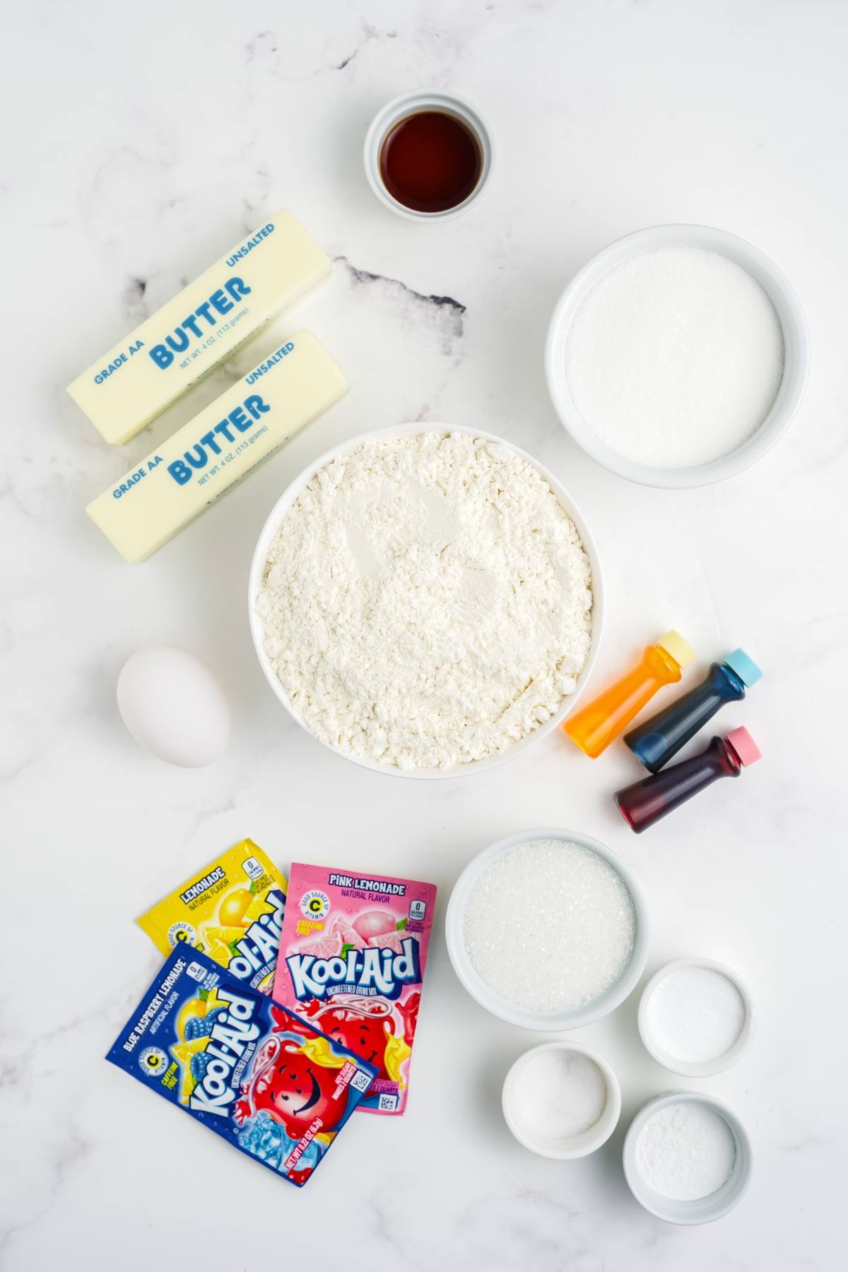Baking ingredients, Kool-Aid packets, food coloring, flour, sugar, butter, egg, and vanilla on a white surface.