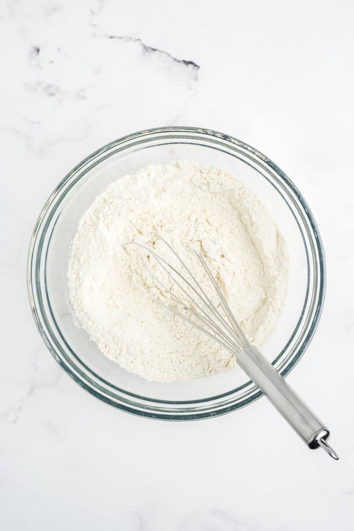 A glass bowl of flour with a metal whisk on a white marble surface.