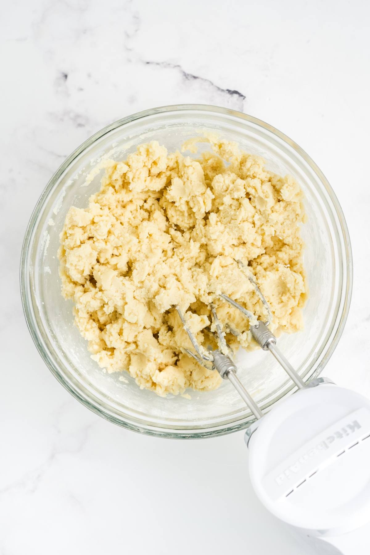A glass bowl of cookie dough being mixed with an electric hand mixer on a marble countertop.