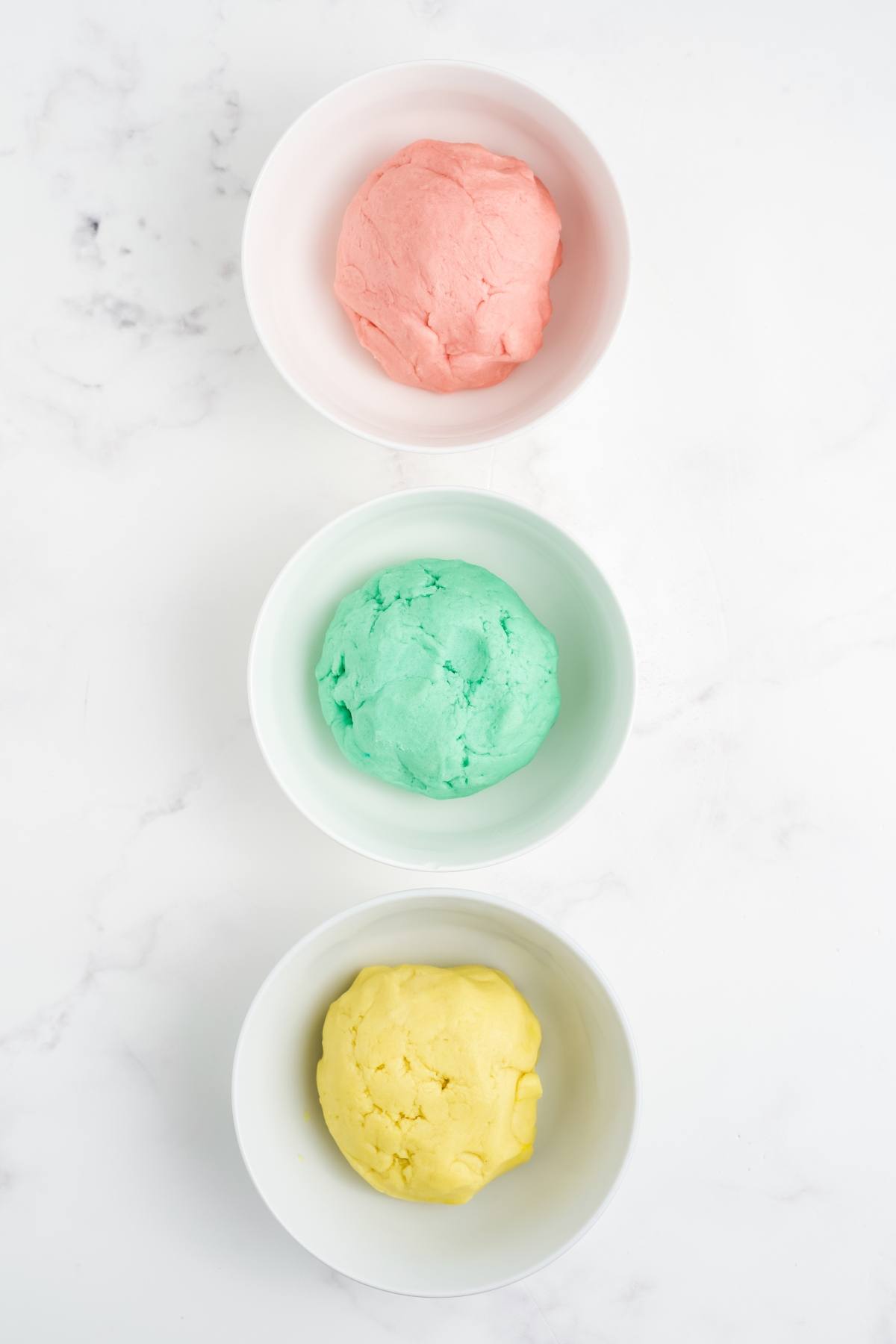 Three white bowls with pink, green, and yellow dough on a white marble surface, arranged vertically.
