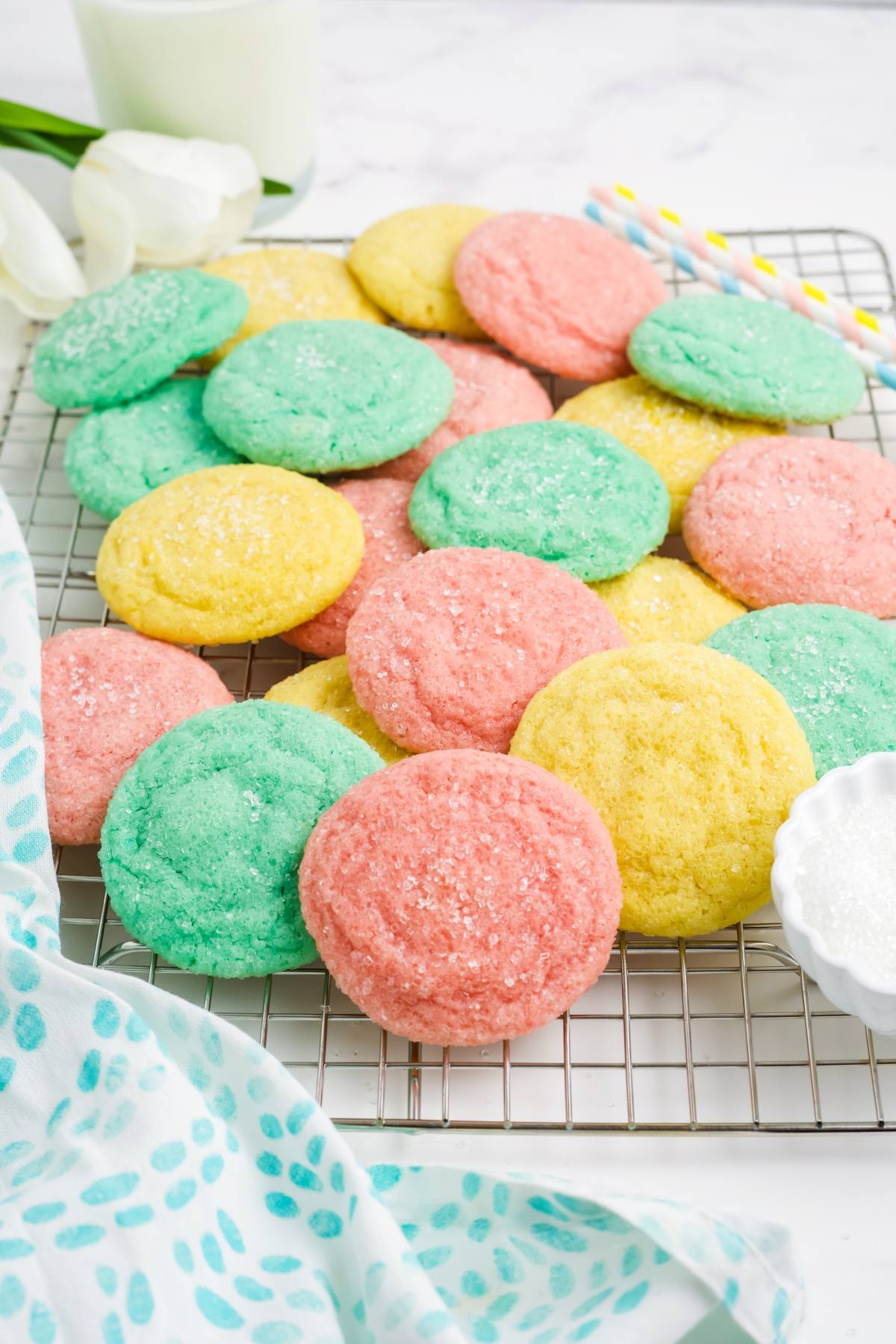 Colorful pink, yellow, and green sugar Kool-Aid cookies on a cooling rack, with a patterned cloth beside them.