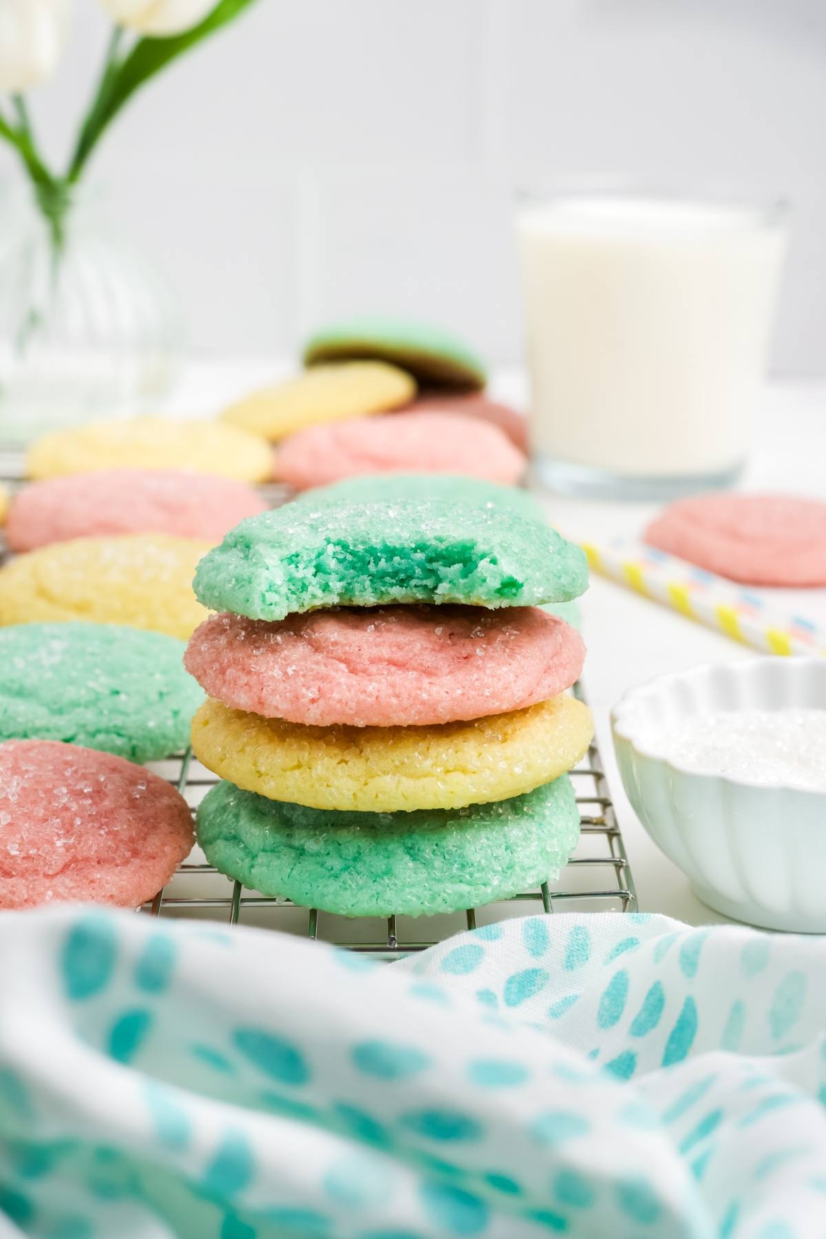 Stack of pastel green, pink, and yellow sugar Kool-Aid  cookies on a cooling rack with a glass of milk nearby.