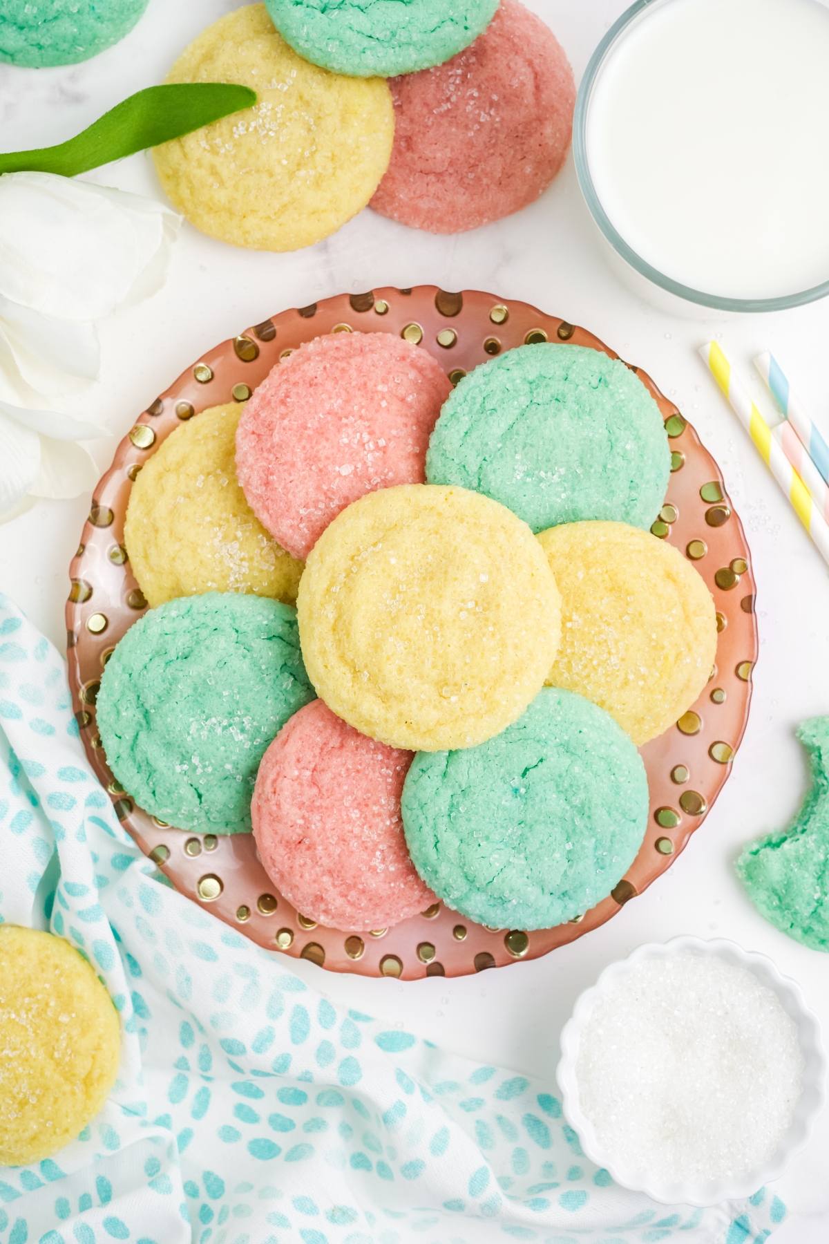 A plate of pastel pink, yellow, and green sugar Kool-Aid cookies next to a glass of milk and a bowl of sugar.