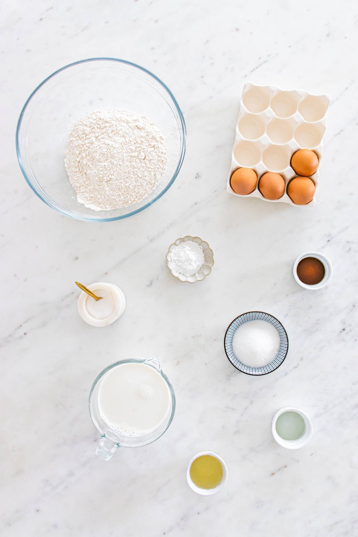 Baking ingredients on a marble surface: flour, eggs, milk, sugar, salt, vanilla, baking powder, oil, and vinegar.
