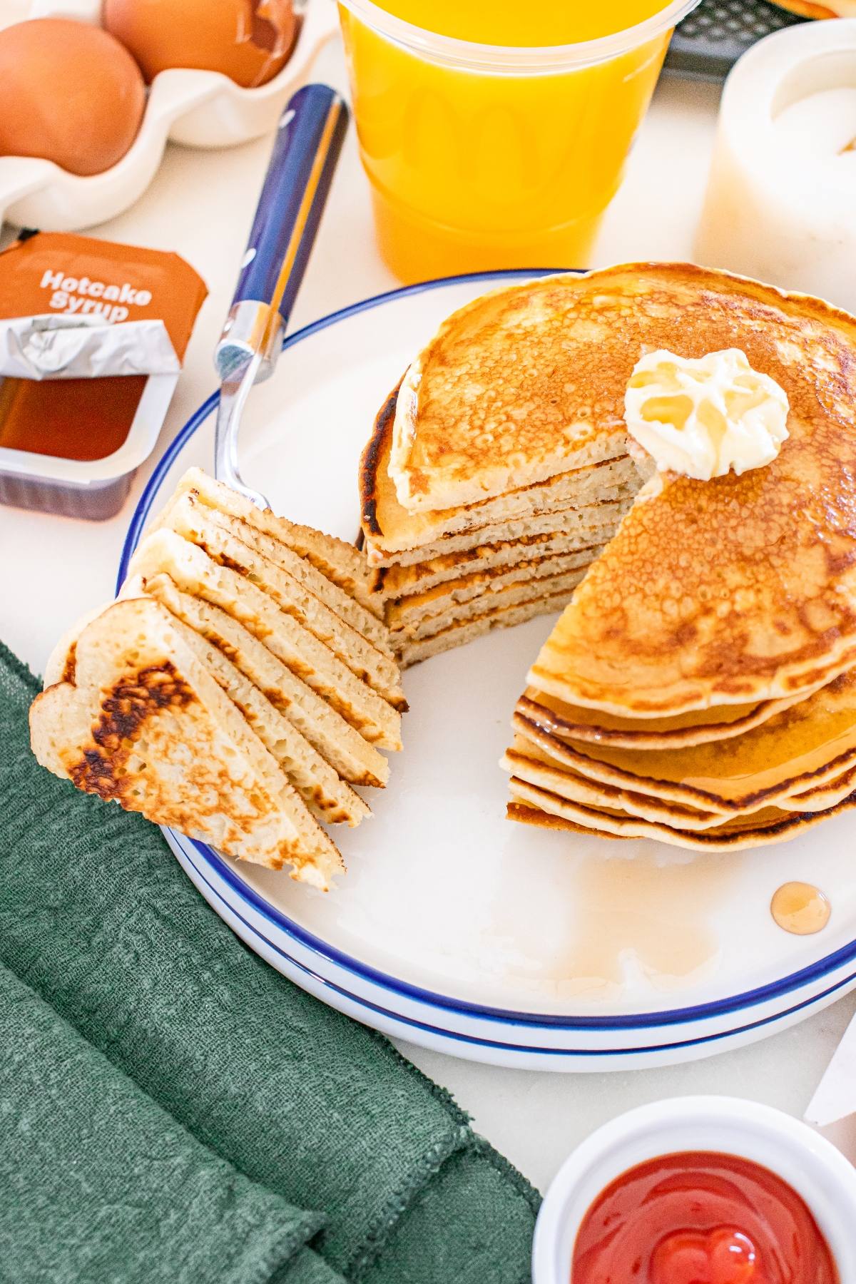 A stack of McDonalds copycat pancakes with butter on top, a fork lifting a slice, surrounded by syrup, eggs, and a yellow cup.