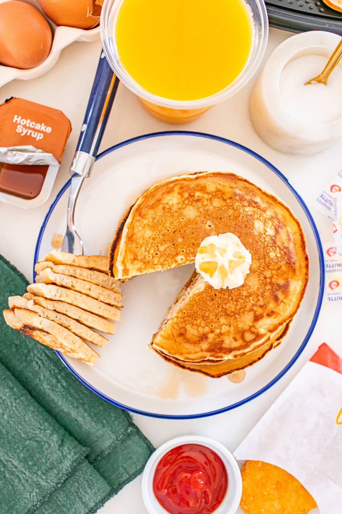 A plate of McDonalds copycat  pancakes with a pat of butter, syrup, orange juice, eggs, and a fork holding sliced pancakes.
