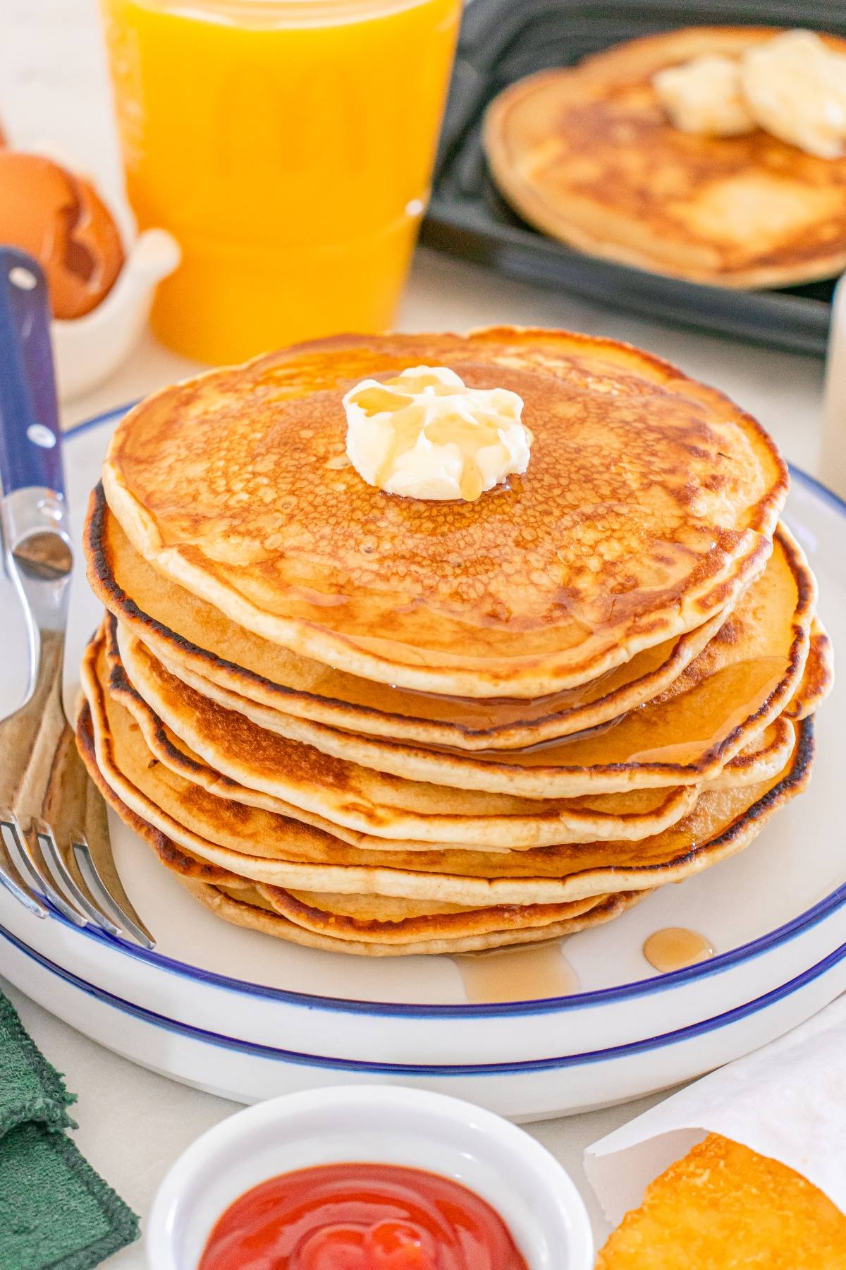 A stack of McDonalds copycat pancakes with butter on top, served on a plate with syrup, fork, and a glass of orange juice.