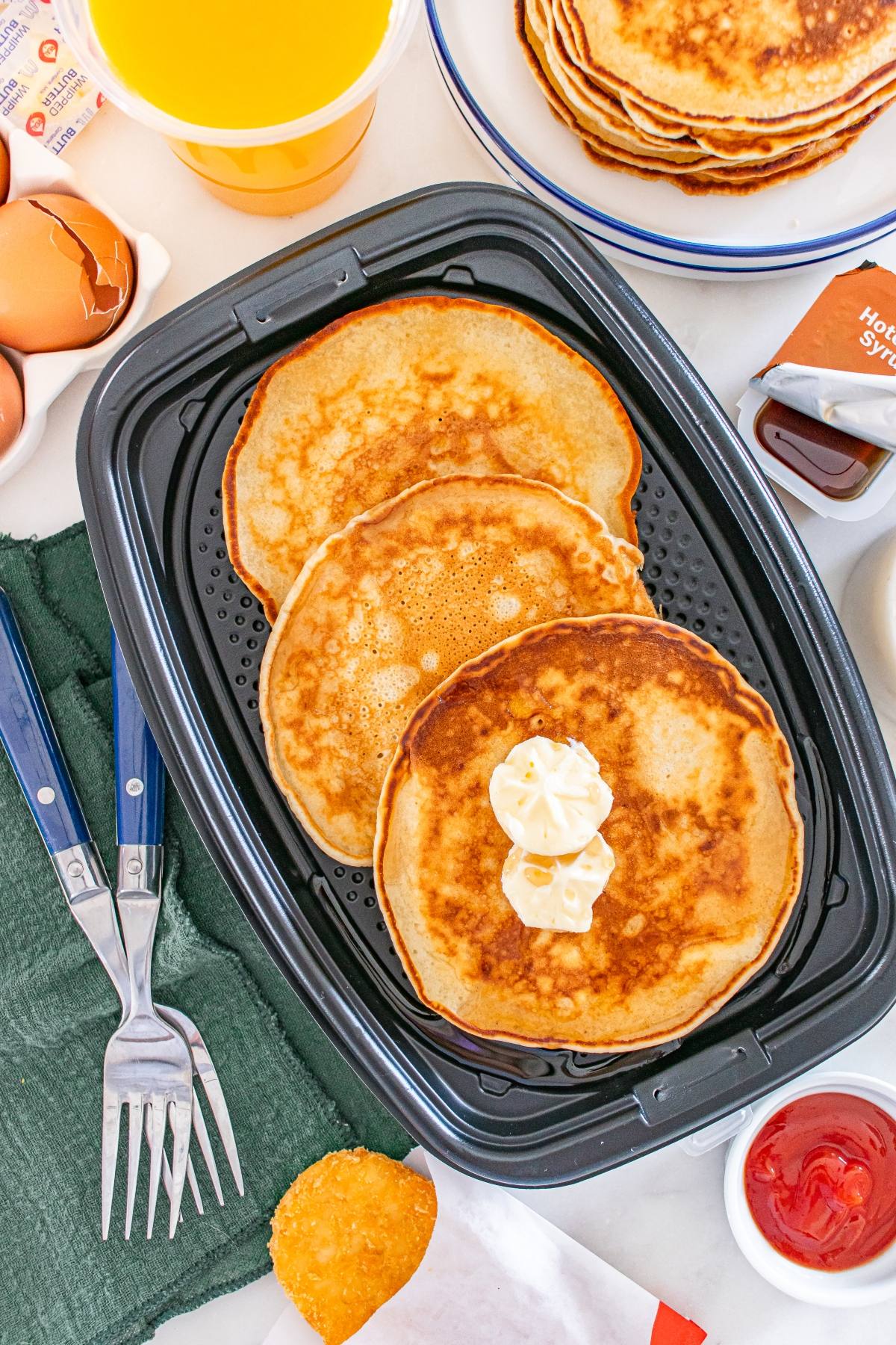 Three McDonald's copycat pancakes with butter on top in an air fryer basket, surrounded by breakfast items and utensils.