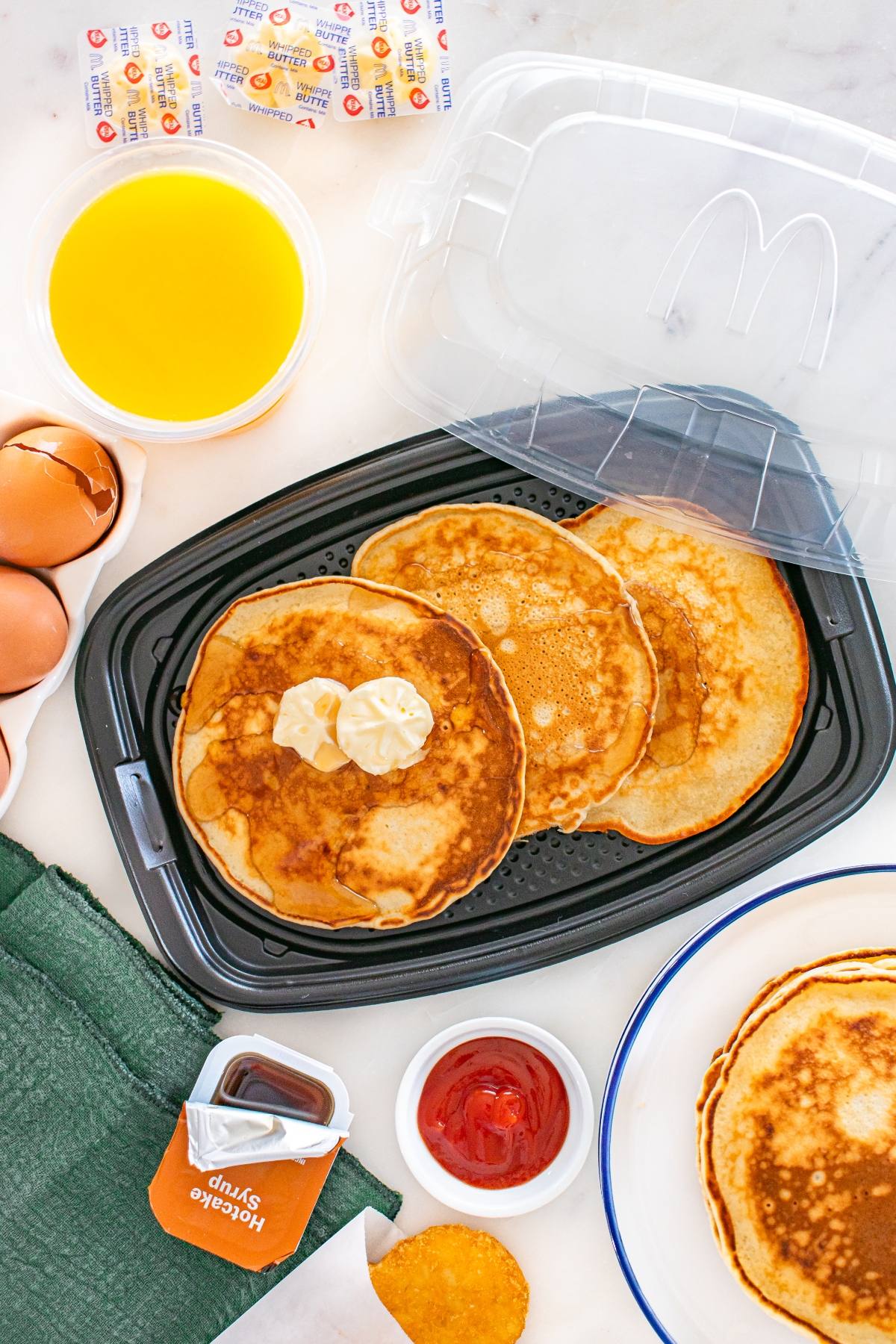 McDonald's copycat pancakes with butter in a plastic tray, eggs, orange juice, syrup, hash brown, and condiments nearby.