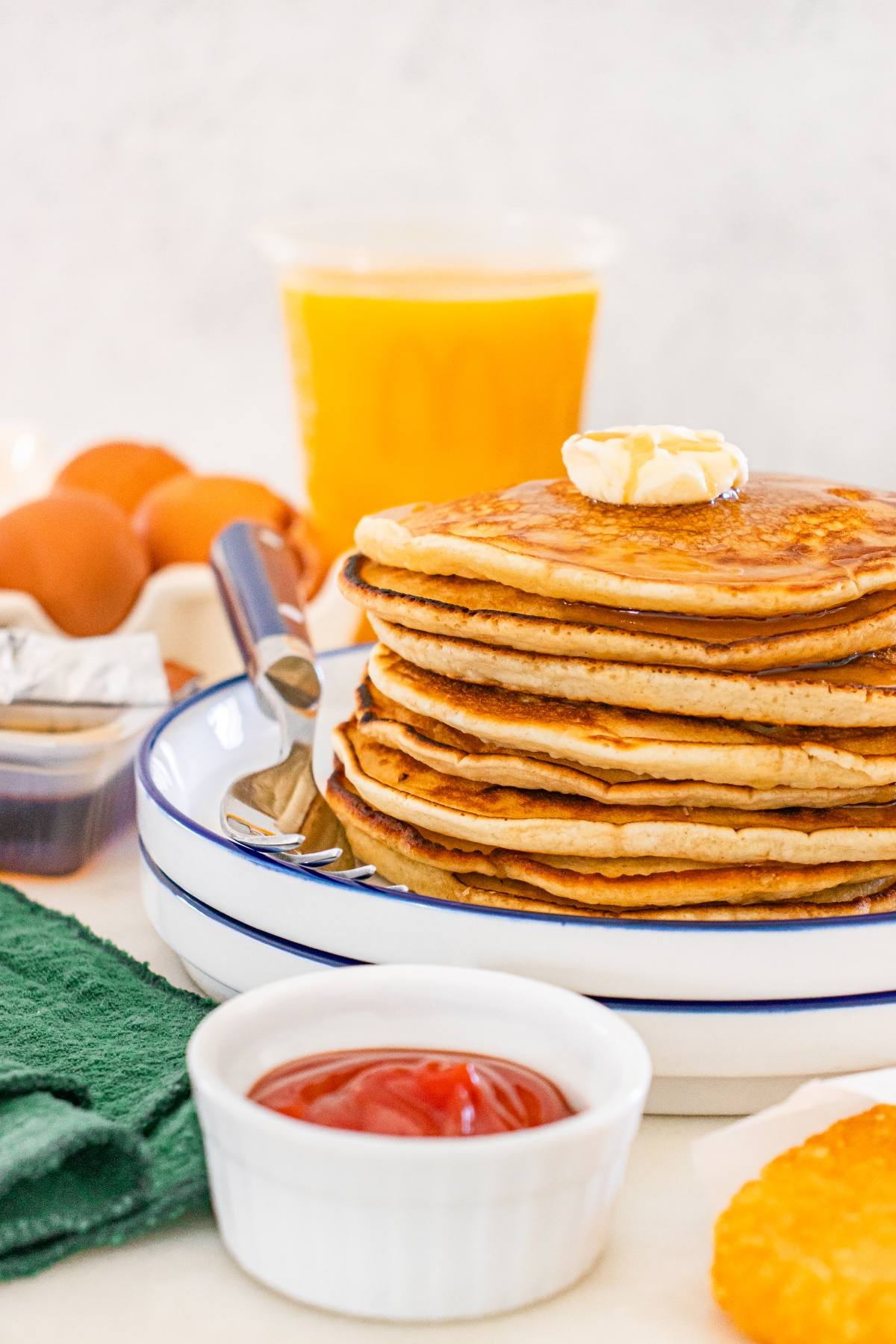 Stack of McDonalds copycat pancakes with butter, syrup, a fork, eggs, orange juice, ketchup, and a hash brown on the table.