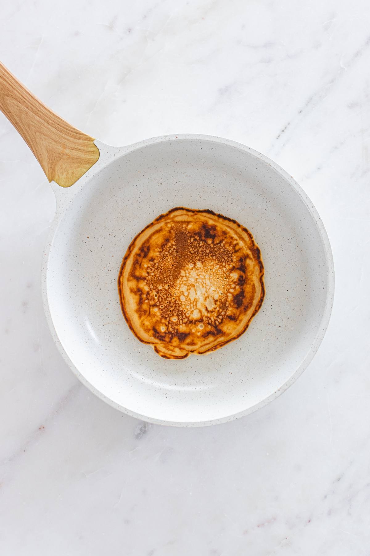 A single pancake cooking in a white frying pan on a marble surface.