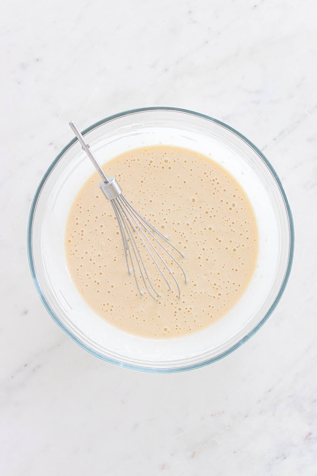 A glass bowl of pancake batter with a whisk, on a white marble surface.