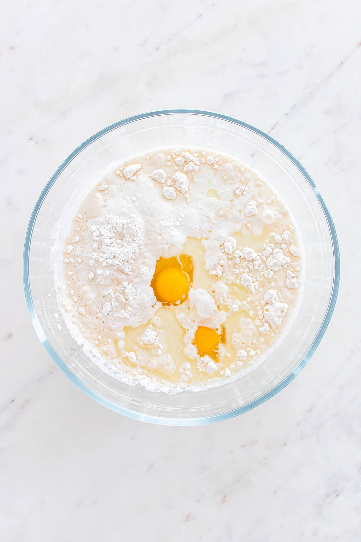 Clear bowl with flour, two eggs, and liquid ingredients on a white marble surface, ready for mixing.