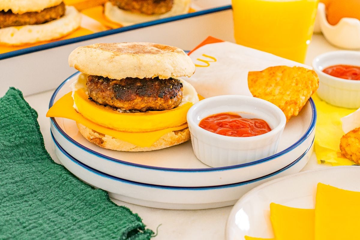 Breakfast sandwich with cheddar, sausage patty, and muffin, served with ketchup and a hash brown on a plate.