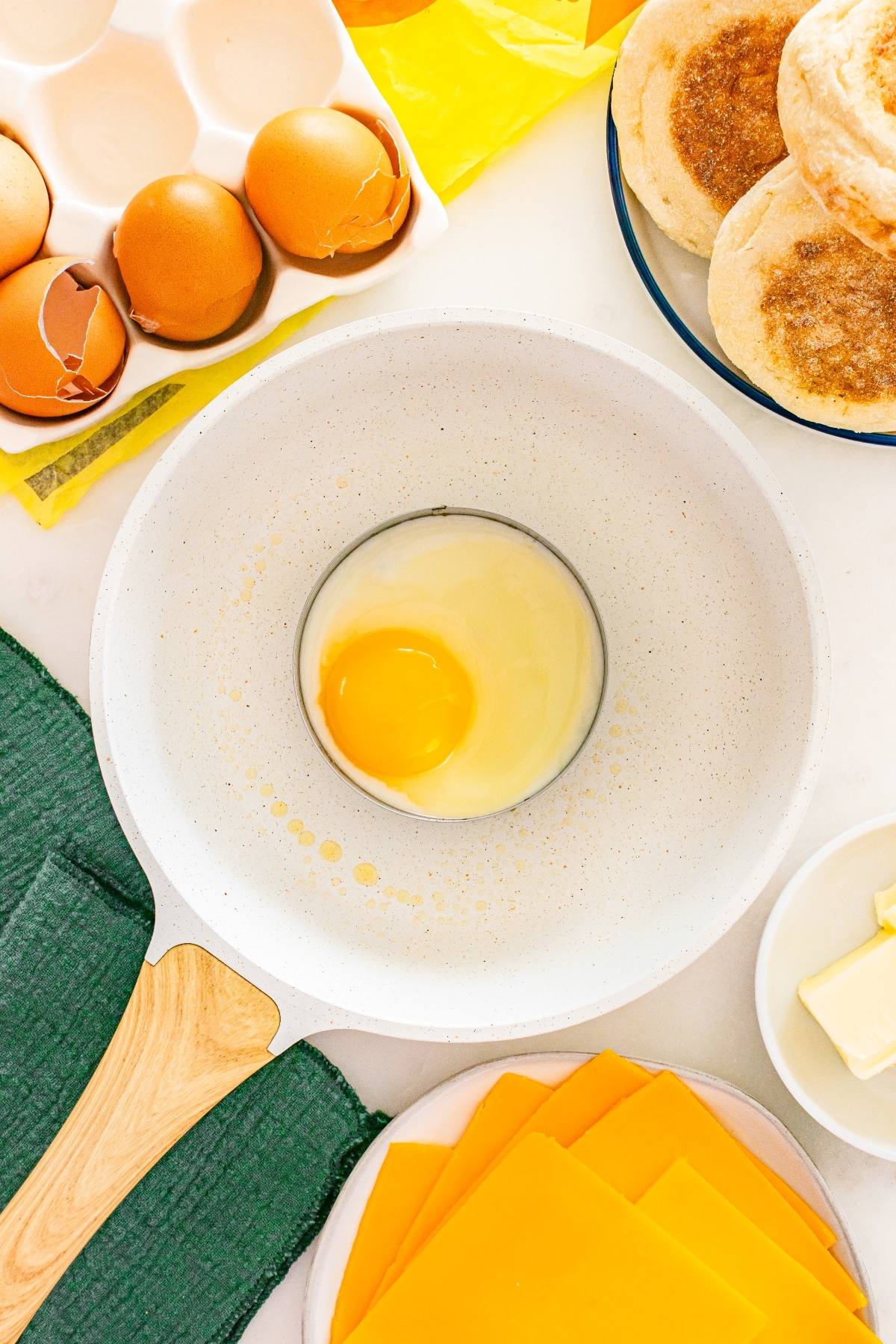 Overhead view of a cracked egg in a bowl, with eggshells, muffins, cheese slices, and butter nearby.