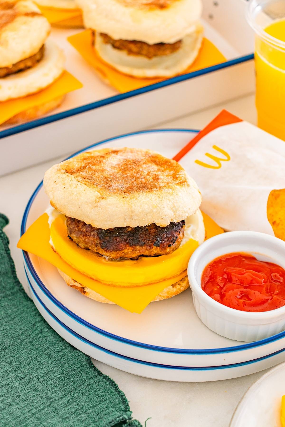 Breakfast sandwich with sausage and cheese on an english muffin, with ketchup and orange juice on the side.