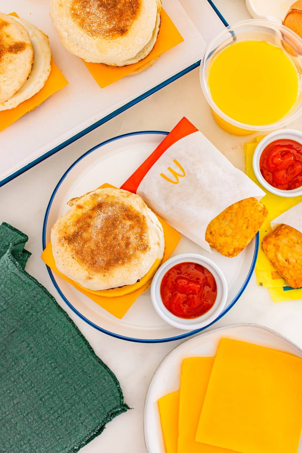 Breakfast sandwich with cheese, hash brown, ketchup, and orange juice on a white table with green napkin.