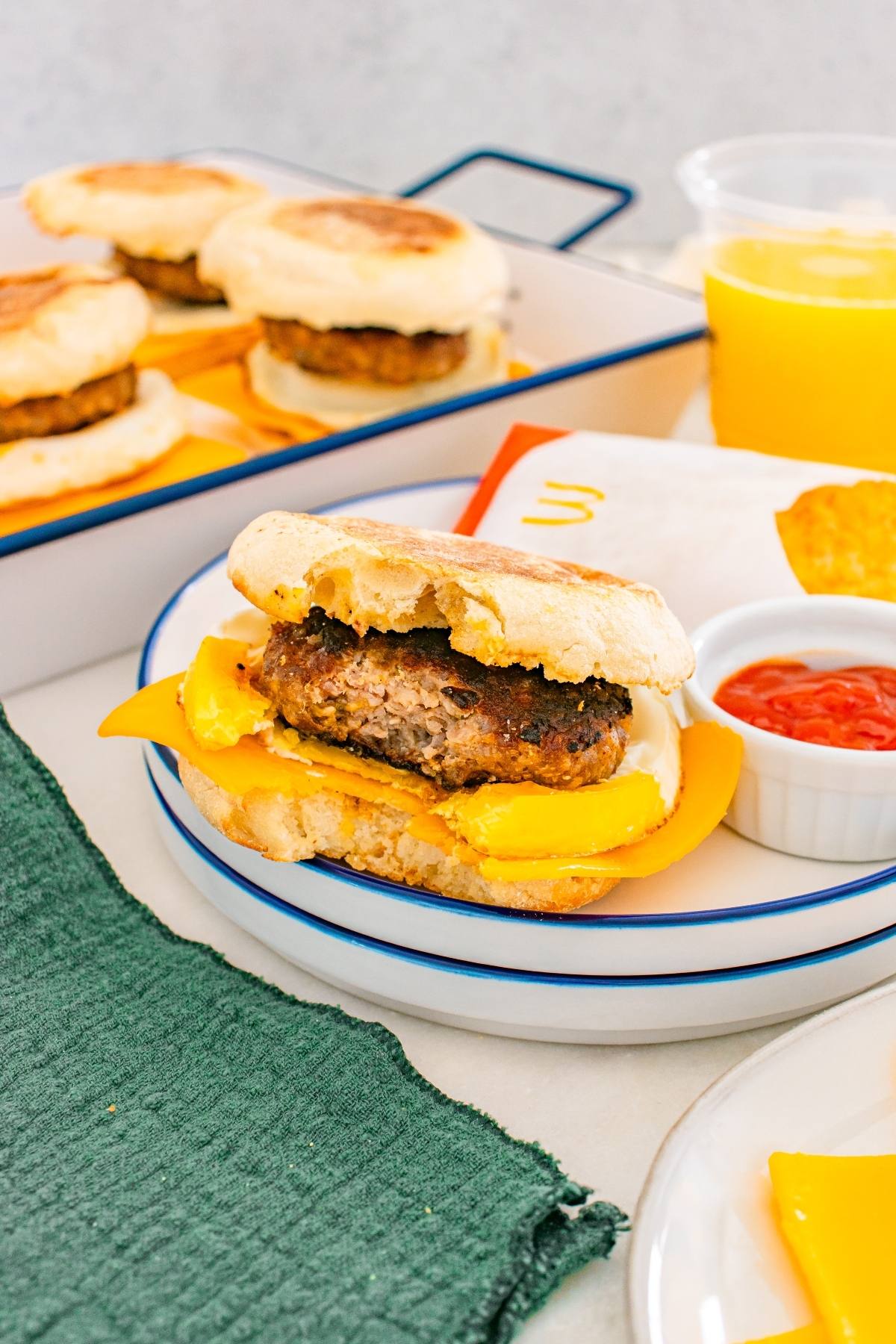 A breakfast sandwich with cheese and patty on an english muffin, with orange juice and ketchup nearby.
