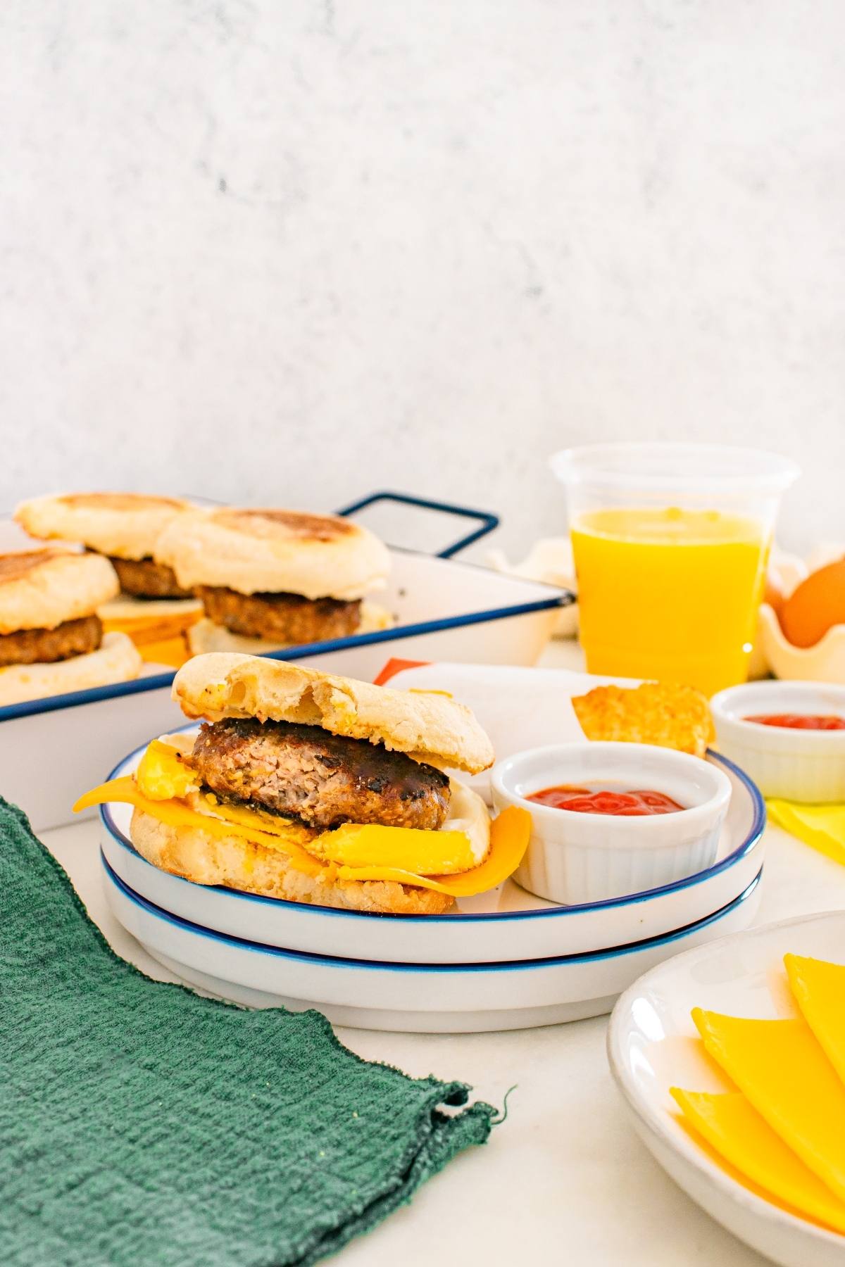 Breakfast sandwich with egg, cheese, and sausage on a plate, with ketchup, orange juice, and muffins nearby.