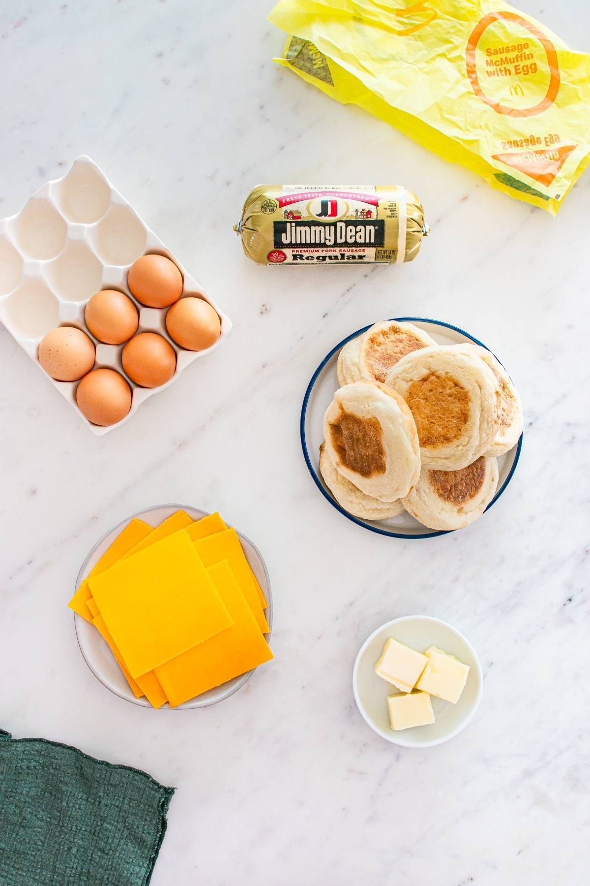Eggs, sausage, english muffins, cheddar cheese slices, and butter arranged on a white marble countertop.