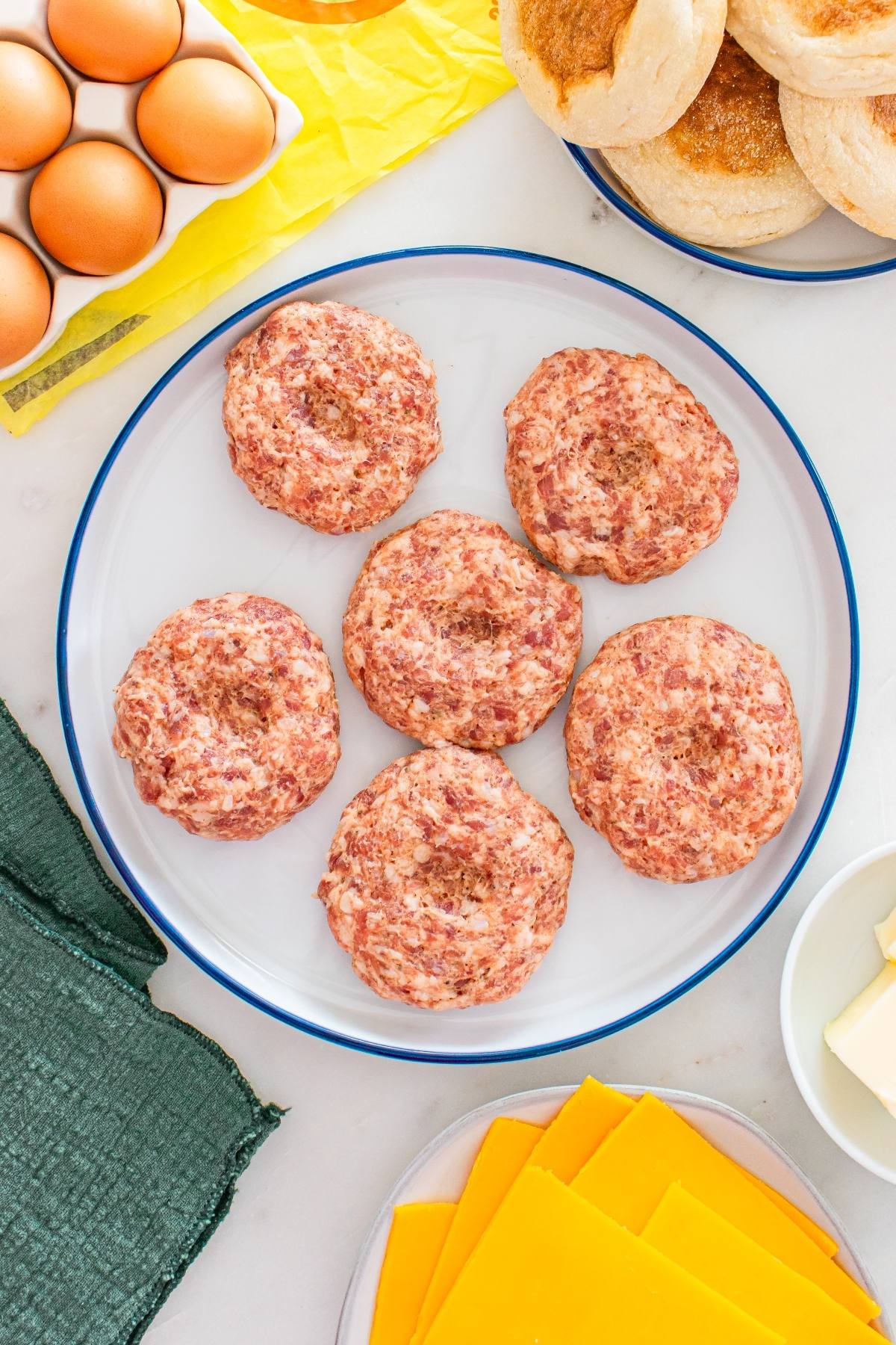 Six raw sausage patties on a white plate, surrounded by eggs, cheese slices, butter, and english muffins.